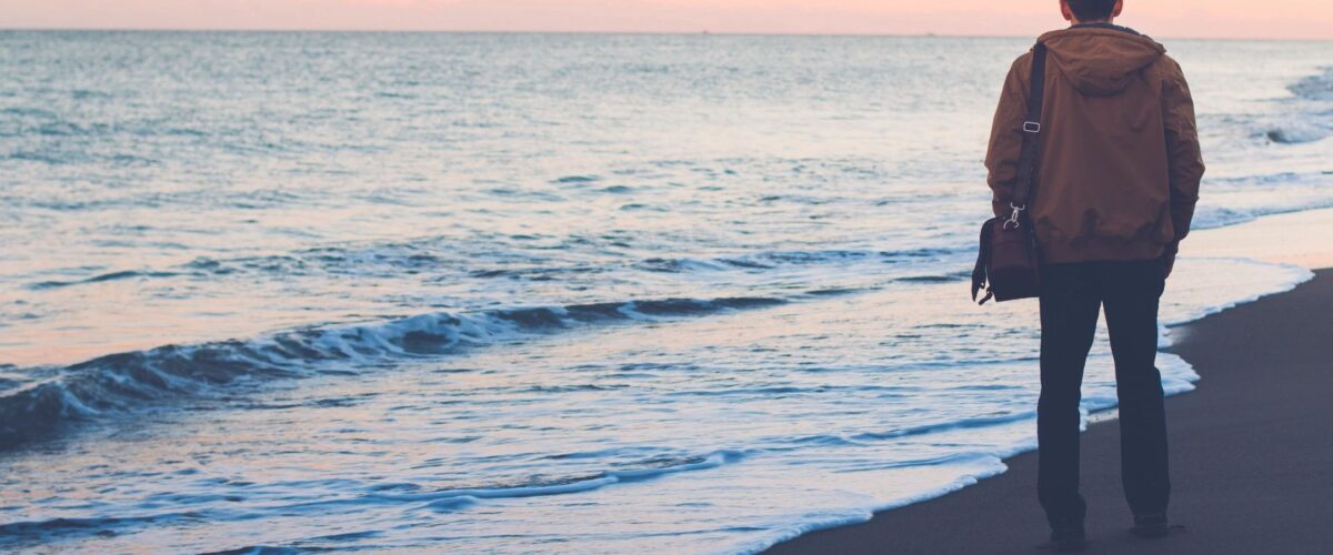 A man standing near the beach at sunset with a laptop bag on his shoulder, reflecting on a simple life, balance, and calm away from work pressure.