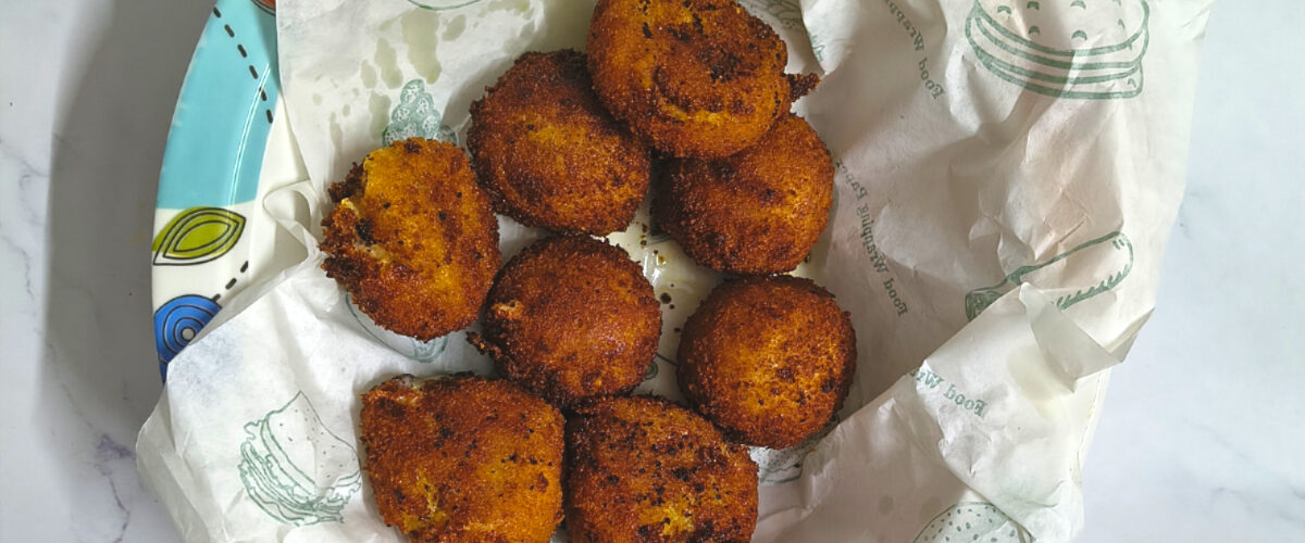 Homemade potato cheese balls served on a plate lined with paper, crispy and golden brown, photographed on a light marble surface with yellow tulips.