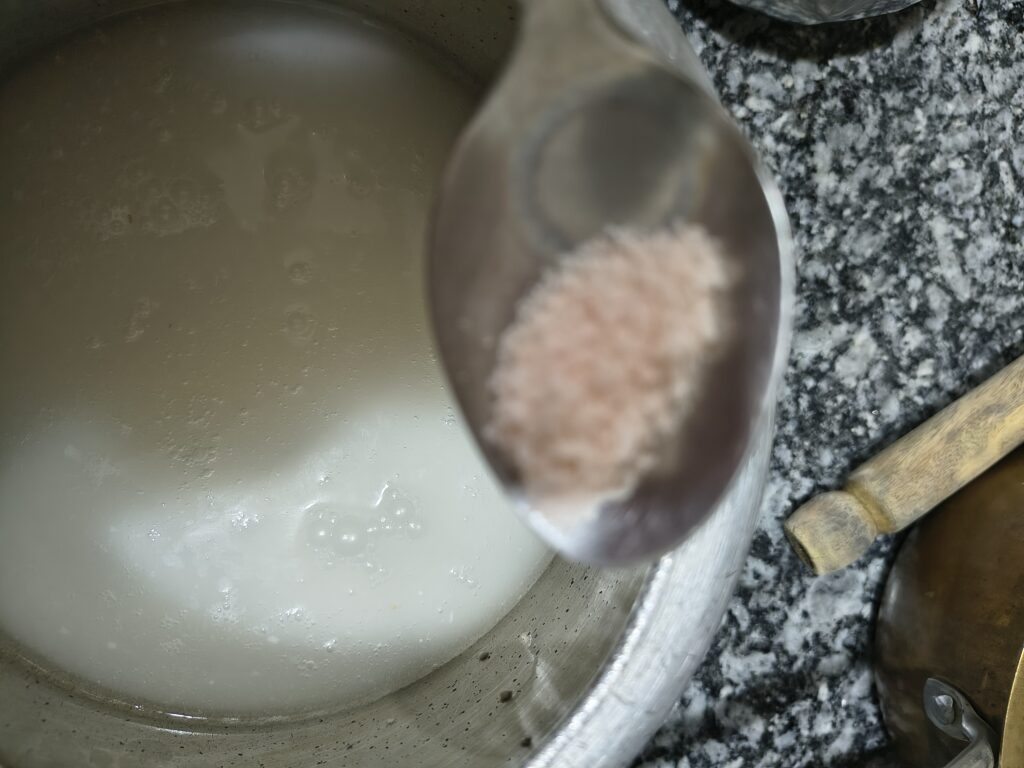 A close-up spoon holding pink rock salt above a pot containing a white mixture of barley water and buttermilk.