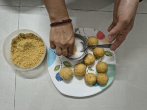 Hands dipping a potato cheese ball into slurry and coating it with breadcrumbs before frying, with several prepared potato balls on a plate.