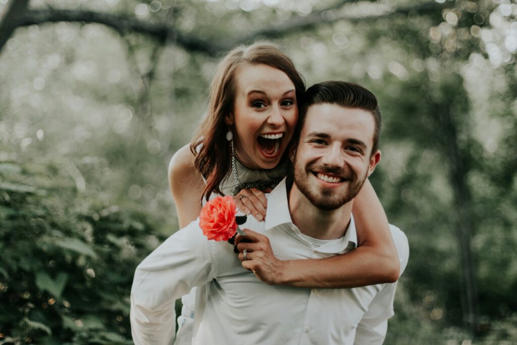 A happy young woman with a big smile riding piggyback on a smiling man holding a pink flower, photographed outdoors in a lush green forest setting.