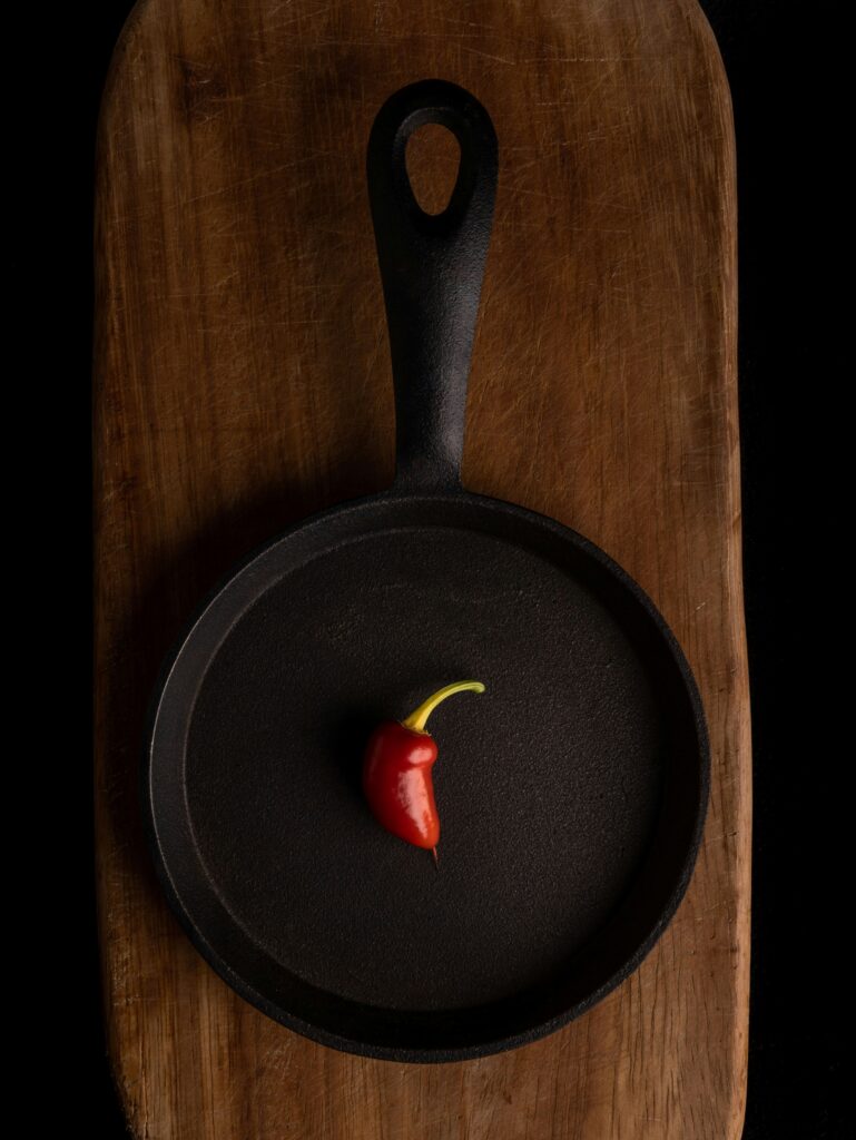 A seasoned black cast iron skillet containing a single red chili pepper resting on a wooden cutting board, representing minimalist kitchen essentials.