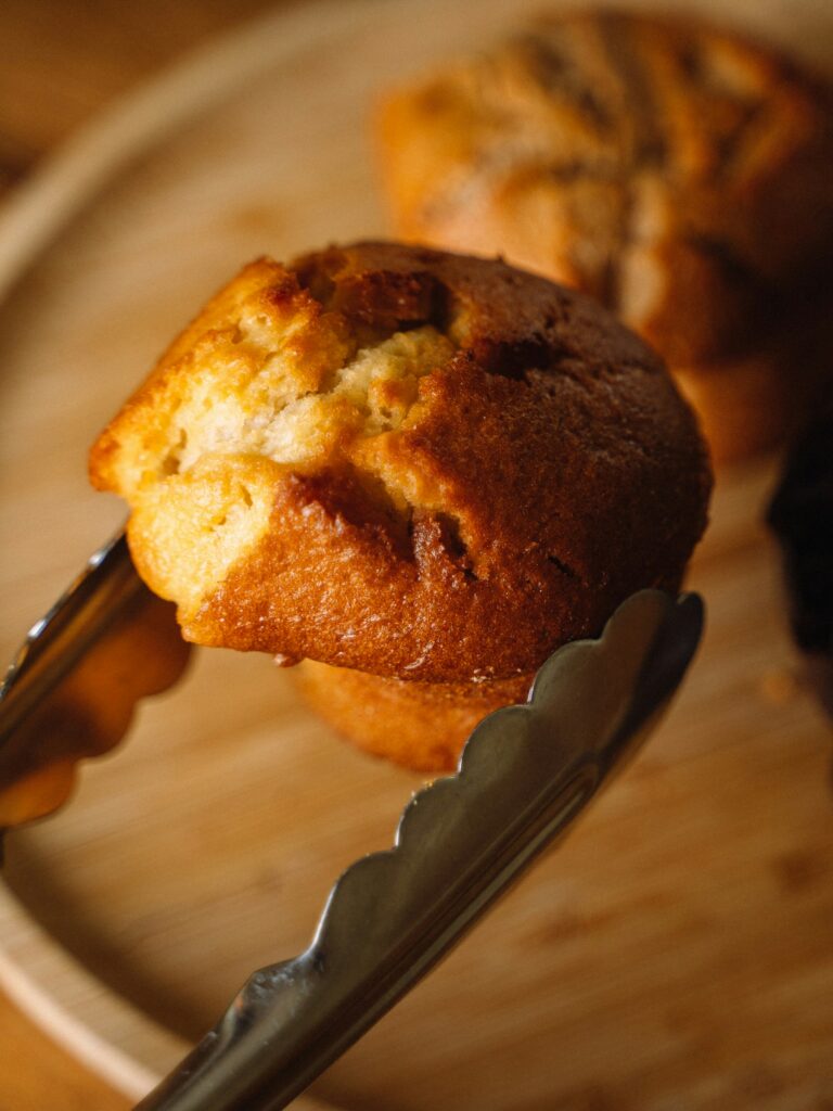  close-up shot of stainless steel kitchen tongs holding a freshly baked golden muffin over a wooden serving tray.