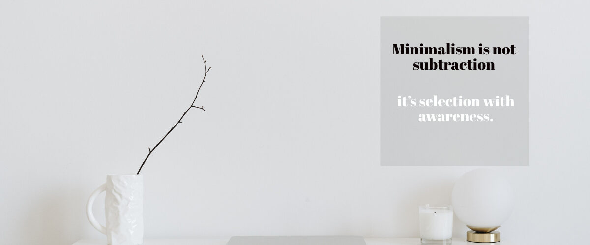 A clean, white minimalist desk setup featuring a closed laptop, a textured white mug holding a single bare branch, a small candle, and a round globe lamp against a white wall with an overlay quote about minimalism.