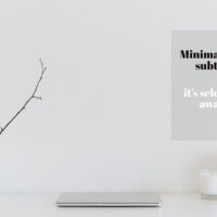 A clean, white minimalist desk setup featuring a closed laptop, a textured white mug holding a single bare branch, a small candle, and a round globe lamp against a white wall with an overlay quote about minimalism.