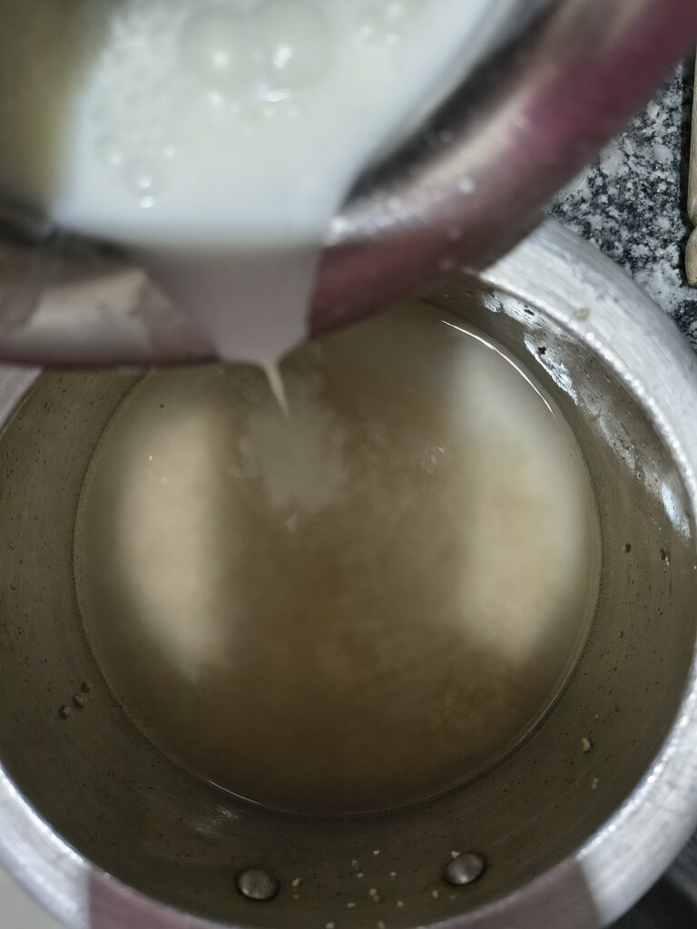 Fresh white buttermilk being poured from one metal container into an aluminum pot filled with cooked barley water.