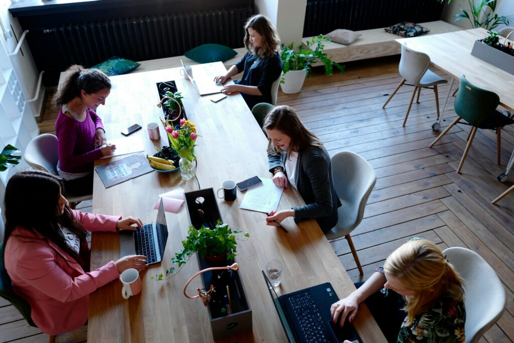 Four women working together at a long wooden table in a bright, plant-filled coworking space, using laptops, writing notes, and drinking coffee.