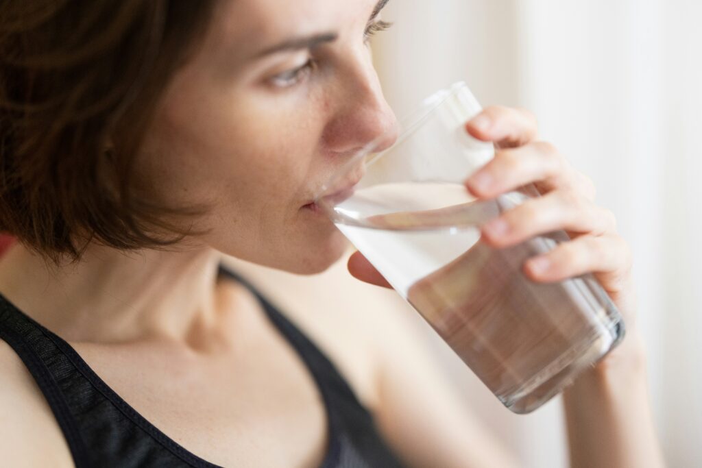 A young girl tilting her head back slightly while drinking clean, refreshing water from a transparent glass.